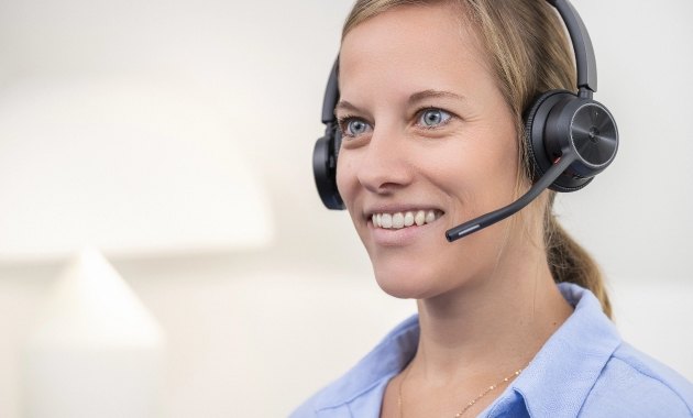 team_hotline Person mit braunen Haaren, hellblauem Hemd und schwarzem Headset vor hellem Hintergrund.